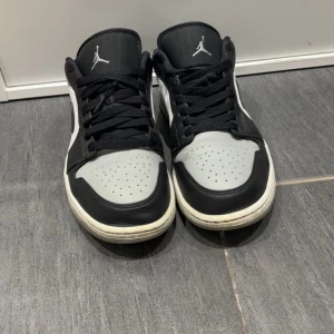 Nike Air Jordan 1 Low svart/grå - Nike Air Jordan 1 Low sneakers i svart, grått och vitt med klassisk Jordan-logga på hälen och tungan. Skorna har snörning, perforerad tå och platt sula. Snygg kontrast mellan panelerna och ikonisk Swoosh på sidan. Perfekt för dig som gillar streetwear och basketstil.