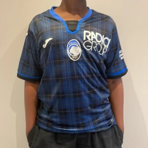 Atalanta matchtröja Holm #3 Joma - Säljer en officiell Atalanta Bergamo fotbollströja från Joma, med spelarnamn Holm och nummer 3 på ryggen. Tröjan har ett blått och svart rutigt mönster, korta ärmar och tryck för Christmas Match 2023. Materialet är lätt och andas, perfekt för match eller träning.