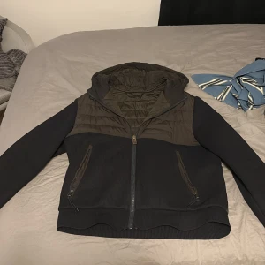 Svart och mörkgrön pufferjacka BOSS - Snygg pufferjacka från BOSS i svart och mörkgrönt med huva och quiltad design. Jackan har ribbade ärmar och nederkant, två dragkedjeförsedda fickor och BOSS-logga på ärmen. Materialet är polyester och stickad textil, perfekt för höst och vinter.