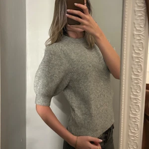 Grå stickad puffärm tröja - Säljer en grå stickad tröja med puffärmar och ribbad kant vid midja och ärmslut. Tröjan har rund hals och korta ärmar som ger en cool och trendig siluett. Perfekt att styla med jeans eller kjol för en chill look.