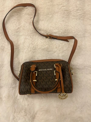 Michael Kors brun axelväska - Snygg axelväska från Michael Kors i brunt med MK-logga över hela väskan. Väskan har detaljer i ljusbrunt skinn, guldfärgade metalldetaljer och dragkedja upptill. Justerbar axelrem och dekorativ MK-berlock. Perfekt storlek för det viktigaste.