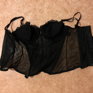 Svart spetskorsett från H&M - Svart korsett från H&M med genomskinliga meshpartier och detaljerad spets framtill och på kuporna. Axelbanden är justerbara och modellen är croppad. Perfekt för att skapa en edgy look med snygga detaljer.