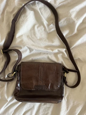 Brun axelväska i skinn från Springfield - Snygg brun axelväska från Springfield i äkta skinn med justerbar rem och praktisk dragkedja på baksidan. Perfekt storlek för det viktigaste och stilren design med diskreta sömmar och metallspännen i mässing.