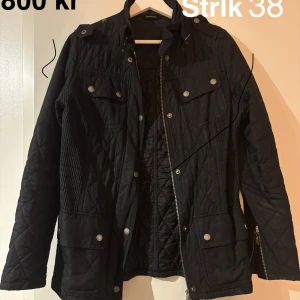 Svart quiltad jacka från Barbour - Quiltad svart jacka från Barbour med snygga silverfärgade knappar och dragkedja. Jackan har flera fickor med lock, hög krage och coola detaljer som axelklaffar. Perfekt för dig som gillar stilren och funktionell design.