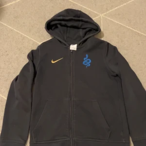 Svart Nike hoodie med blått tryck - Svart hoodie från Nike med dragkedja, huva och fickor framtill. På bröstet finns ett blått tryck med orm och en gul Nike-logga. Perfekt för chill eller träning, riktigt skön och stilren. Materialet är mjukt och känns som bomull.