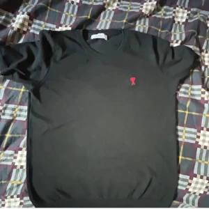 Svart stickad tröja från Ami Paris - Svart stickad tröja från Ami Paris med klassisk rund hals och korta ärmar. På bröstet finns Ami:s ikoniska röda hjärta med A broderat. Tröjan har ribbade muddar vid hals och ärmslut, vilket ger en clean och stilren look.