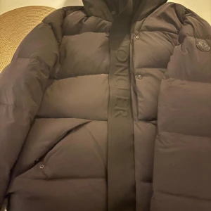 Svart Moncler vinter jacka  - Säljer en svart pufferjacka från Moncler med huva och tydlig logga på bröstet. Jackan har breda, quiltade sektioner och en snygg Moncler-detalj längs dragkedjan. Perfekt för kalla dagar och har praktiska fickor med knappstängning.