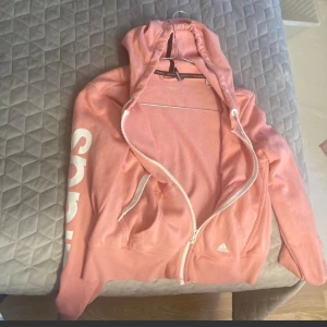 Rosa hoodie från Adidas med dragkedja - Snygg rosa hoodie från Adidas med vit dragkedja och snörning i huvan. Stor Adidas-logga i vitt längs ena ärmen och liten logga på bröstet. Tillverkad i mjuk bomull med Climalite-teknologi som andas. Perfekt för chill eller träning.