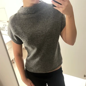 Grå stickad polotröja kortärmad - Säljer en grå stickad polotröja med korta ärmar och hög krage. Tröjan har en enkel och clean look, perfekt för dig som gillar minimalistisk stil. Materialet är mjukt och skönt mot huden och passformen är normal.