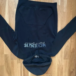 McKenzie hoodie  - Svart hoodie från McKenzie med broderad logga i silver på bröstet. Tröjan har huva med snörning och en stor magficka framtill. Långärmad och tillverkad i mjuk bomullsmix som är skön att ha på sig. Perfekt för chill eller streetwear.