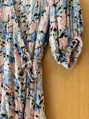 Blommig omlottklänning Gina Tricot - Säljer en kort omlottklänning från Gina Tricot i storlek 36. Klänningen har blommigt mönster i ljusa toner av rosa, blått, grönt och svart. Den har korta ärmar, knyt i midjan och är tillverkad i mjuk viskos. Perfekt för dig som gillar romantiska och färgglada plagg. Blommig omlottklänning med knytband och puffärm. 