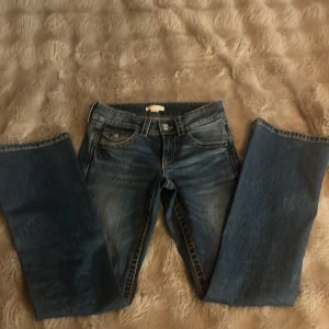 Blå bootcut jeans från Gina Tricot - Snygga blå jeans från Gina Tricot i bootcut-modell med kontrastsömmar och coola fickdetaljer med knapp bak. Klassisk femficksdesign och bälteshällor. Perfekt för dig som gillar en lite retro vibe och vill ha jeans med lite extra attityd.
