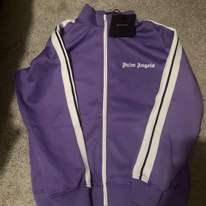 Palm angels kofta Lila  - Palm Angels lila track jacket med vita och svarta ränder längs ärmarna. Jackan har dragkedja framtill, ribbade muddar och klassisk logga på bröstet. Tillverkad i 100% polyester för en sportig vibe. Perfekt för dig som gillar streetwear och vill sticka ut.