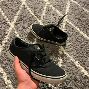 Svarta Vans sneakers med vit sula - Klassiska svarta Vans sneakers i canvas med vit sula och bruna gummisulor undertill. Skorna har snörning och låg profil, perfekt för en avslappnad streetstil. Diskret Vans-logga på sidan och platt sula för skön känsla.
