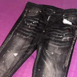 Svarta Dsquared2 jeans med färgstänk - Säljer ett par svarta jeans från Dsquared2 med coola slitningar och färgstänk i vitt och rött. Jeansen har en slim passform, klassiska fem fickor och snygg Dsquared2-lapp bak. Tillverkade i Italien i stretchig bomull för skön känsla. Ny pris 4800 mitt pris 4000 priset kan sänkas 
