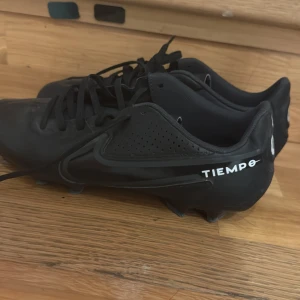 Nike Tiempo svarta fotbollsskor - Säljer ett par svarta Nike Tiempo fotbollsskor med klassisk design och snörning. Skorna har dobbar för bra grepp på planen och är tillverkade i syntetmaterial med diskret Nike-logga på sidan. Perfekta för dig som vill ha stilrena och funktionella skor till matchen.
