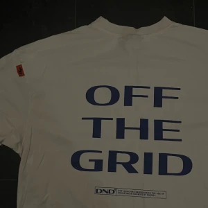 Pegador t shirt - Säljer en vit t-shirt från DND med stor blå text 'OFF THE GRID' på ryggen och mindre tryck fram. T-shirten har korta ärmar och en klassisk rund hals. Materialet är mjuk bomull och det finns en orange detalj på ena ärmen. Perfekt för dig som gillar streetwear.
