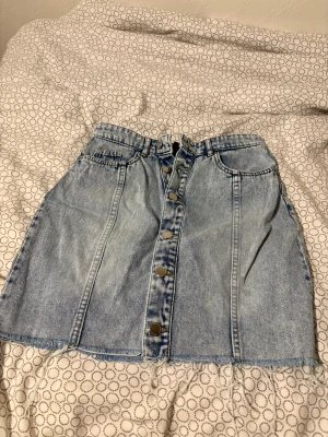Blå jeanskjol med knappar - Säljer en snygg ljusblå jeanskjol med fransad nederkant och knappar hela vägen framtill. Kjolen har klassiska fickor och en cool, avslappnad vibe som passar perfekt till sneakers eller boots. Perfekt för dig som gillar denim och vill ha en trendig look.