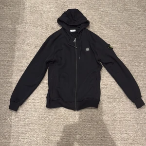 Svart hoodie från Stone Island L - Svart hoodie från Stone Island i storlek L, tillverkad i 100% bomull. Tröjan har huva med snörning, dragkedja framtill och klassisk Stone Island patch på vänster ärm. Snygg och enkel design med ribbade muddar och två fickor framtill.