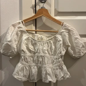 Vit puffärmad blus från H&M - Supersöt vit blus från H&M med puffiga korta ärmar och smockad midja. Toppen har volang nertill och en luftig känsla. Perfekt för dig som gillar en feminin och gullig stil. Är från 10-11.