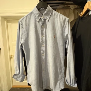 Blå slim fit skjorta från Polo Ralph Lauren - Snygg ljusblå skjorta från Polo Ralph Lauren i slim fit-modell. Klassisk button-down krage, broderad logga på bröstet och knappar hela vägen. Tillverkad i stretchig bomull för extra komfort. Perfekt för dig som gillar stilrena och tidlösa plagg.