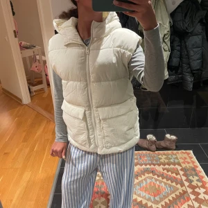 Vit dunväst med huva och fickor - Säljer en fluffig vit dunväst med stor huva och två rejäla fickor framtill. Västen har dragkedja hela vägen och är quiltad för extra värme. Perfekt att slänga över en hoodie när det är kyligt ute. Enkel och clean design som funkar till många stilar. Stl 164 barn men lite stor på mig som är xs. Använd en gång.
