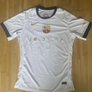 Barcelona Lamine Yamal vit matchtröja - Säljer en vit FC Barcelona fotbollströja från Nike med Lamine Yamal och nummer 10 på ryggen. Tröjan har korta ärmar med svarta muddar, guldfärgade detaljer och klubbmärke på bröstet. Materialet är lätt och andas, perfekt för match eller träning.