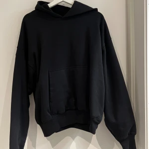 Svart oversized YZY hoodie - Svart hoodie från YZY med oversized passform och stor huva. Tröjan har en klassisk känguruficka framtill och ribbade muddar vid ärmslut och nederkant. Perfekt för chill dagar och streetwear-stil. 