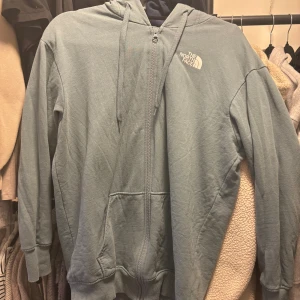 Blå/Grön hoodie från The North Face - Snygg blå hoodie från The North Face med dragkedja och huva. Tröjan har snörning vid halsen, ribbade muddar och fickor framtill. 