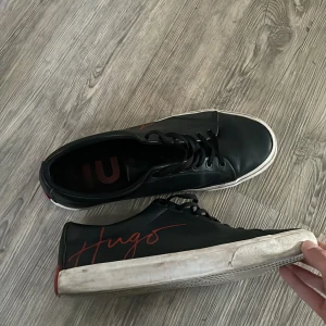 Svarta Hugo sneakers med röd logga - Snygga svarta sneakers från Hugo med röd logga på sidan och röd detalj på hälen. Skorna har snörning, rund tå och vit platt sula med Hugo-mönster undertill. Tillverkade i skinn för en clean och modern look, perfekt för dig som gillar stilrena detaljer. Pris ej hugget i sten så hör av er vid minsta fundering✅👍