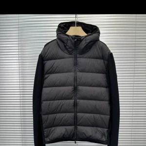 Svart Moncler pufferjacka med huva - Svart pufferjacka från Moncler med stickade ärmar och huva. Jackan har quiltad framsida i syntetmaterial och dragkedja hela vägen. Moncler-logga på ärmen. Perfekt för dig som gillar stilren och sportig look.