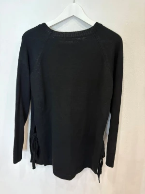 Svart stickad tröja med snörning - Svart stickad tröja med rund hals och långa ärmar. Tröjan har snygg snörningsdetalj på sidorna som ger en cool touch. Perfekt för dig som gillar enkel stil med en twist. Passar dig som vill ha en basic men ändå unik tröja.