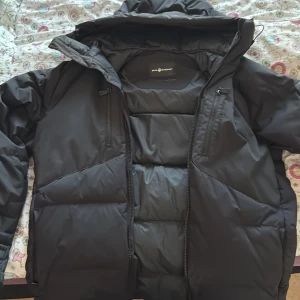 Svart pufferjacka Sail Antarctica - Svart pufferjacka från Sail Antarctica med huva och flera dragkedjor. Jackan har en stilren, quiltad design och är tillverkad i syntetmaterial som håller dig varm. Perfekt nu när de börjar bli kallt. De enda skadorna är att inner drag kedjan inte funkar och patchen har gått sönder men annars är jackan i gott skick, därav de nedsatta priset.