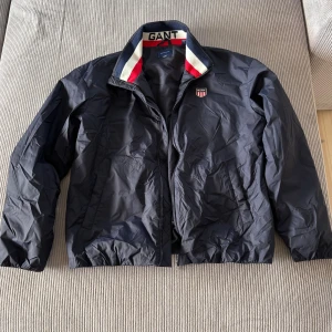 Svart vindjacka från GANT med logga - Svart vindjacka från GANT med coolt broderat märke på bröstet och dragkedja framtill. Kragen har snygga ränder i rött, vitt och marinblått med GANT-text. Jackan har två fickor och elastisk nederkant för skön passform.