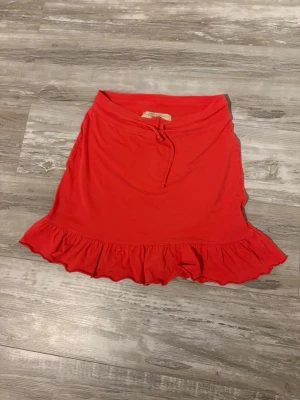 Röd volangkjol med snörning - Snygg röd kortkjol från VERO MODA med volangkant längst ner och snörning i midjan. Kjolen är gjord i mjuk bomull och har en enkel, feminin design som ger en cool vibe. Perfekt för dig som gillar färg och vill sticka ut lite extra.