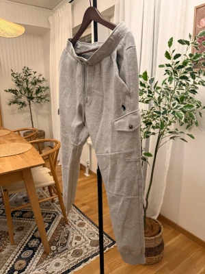 Grå mjukisbyxor från Polo Ralph Lauren - Snygga grå mjukisbyxor från Polo Ralph Lauren med snörning i midjan och praktiska cargofickor med knapp. Byxorna har en relaxed passform och är tillverkade i mjukt bomullsmaterial. Perfekt för chill dagar och streetwear-stil.