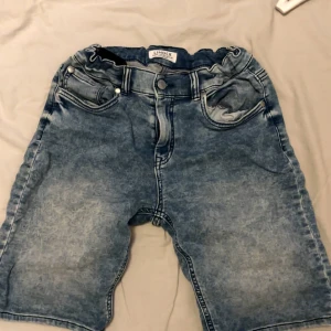 Blå denimshorts från Lindex - Säljer ett par blå denimshorts från Lindex med klassisk femficksdesign och knappstängning. Shortsen har en loose passform och midwaist, med snygga slitningar och tvättad look som ger en cool vibe. Perfekta för varma dagar och chill häng.
