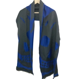 Blå och svart ullhalsduk Alexander McQueen - Snygg halsduk från Alexander McQueen i blått och svart med stort döskallemotiv. Tillverkad i Italien av mjuk ull, mått 35x180 cm. Perfekt accessoar för dig som vill sticka ut med en cool design och exklusiv känsla. Bra skick, några dragna trådar och slitage efter tvätt finns dock.