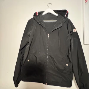 Svart vindjacka från Moncler - Svart vindjacka från Moncler med huva och dragkedja framtill. Jackan har Moncler-logga på vänster ärm och snygga detaljer med röd och vit rand på huvan. Två fickor med knappstängning och justerbar dragsko vid huvan. Perfekt för dig som gillar stilrena och sportiga plagg. 