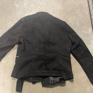 Svart mockajacka från Piazza Italia - Snygg svart mockajacka från Piazza Italia i storlek XL. Jackan har coola detaljer som sned dragkedja, bälte i midjan och dragkedjor vid fickor och ärmslut. Perfekt för dig som gillar en stilren och edgy look.