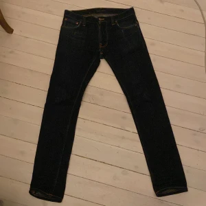 Mörkblå jeans från Nudie Jeans Co - Säljer ett par mörkblå jeans från Nudie Jeans Co i ekologisk bomull. Klassisk femficksmodell med orange kontrastsömmar och Nudie-logga på bakfickan. Jeansen har en rak passform och normal midja, perfekta för en clean och stilren look. W:30 L:32
