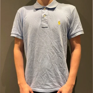 Ljusblå pikétröja från Ralph Lauren - Snygg ljusblå pikétröja från Ralph Lauren med klassisk krage, korta ärmar och två knappar framtill. Ikoniska gula loggan broderad på bröstet. Perfekt för en clean och stilren look.