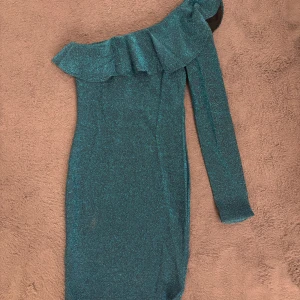 Glittrig grön one shoulder klänning från Nelly - Snygg glittrig grön kortklänning från Nelly i storlek XS. Klänningen har en one shoulder-design med volang upptill och lång ärm på ena sidan. Använd vid endast två tillfällen😊🌸