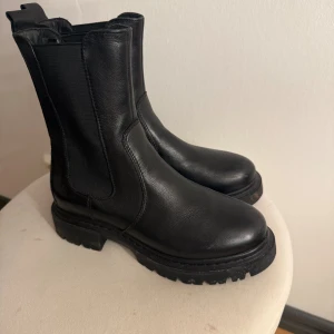 Svarta chunky chelsea boots - Säljer ett par svarta chelsea boots med grov, chunky sula och rund tå. Bootsens ovandel är i slätt skinn och har elastiska paneler på sidorna för enkel på- och avtagning. Perfekta för dig som gillar en edgy och stilren look.
