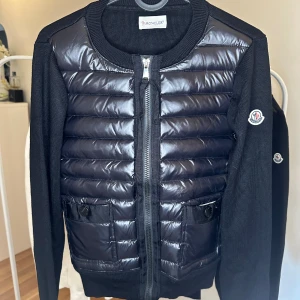 Moncler cardigan  - Snygg svart pufferjacka från Moncler med glansig, quiltad framsida och två stora fickor med knapp. Jackan har långärmade stickade ärmar och rund hals. Dragkedja framtill och Moncler-logga på ärmen. Perfekt för dig som gillar exklusiv streetstyle.