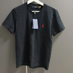 Svart t-shirt från Polo Ralph Lauren - Svart t-shirt från Polo Ralph Lauren med klassisk rund hals och korta ärmar. T-shirten är i mjuk bomull och har den ikoniska röda broderade loggan på bröstet. Perfekt för dig som gillar stilrena och tidlösa plagg med en touch av lyx. 