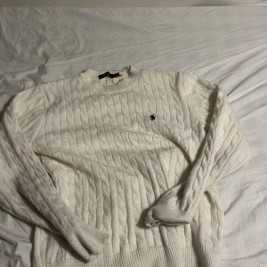 Vit kabelstickad tröja Polo Ralph Lauren - Vit kabelstickad tröja från Polo Ralph Lauren med klassisk rund hals och broderad logga på bröstet. Tröjan har ribbade muddar och är långärmad, perfekt för dig som gillar stilren och tidlös design.Scick 8/10 då den har ett litet hål i sömmen men det går att sy ihop rätt enkelt hör av er vid funderingar