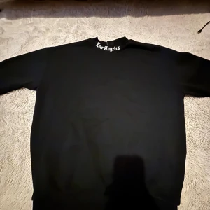 Svart oversized sweatshirt Los Angeles - Svart oversized sweatshirt från Black Squad med stor vit 'Los Angeles'-text på ryggen och mindre text vid halsen. Tröjan har lång ärm och är gjord i mjuk bomull, perfekt för en chill streetwear-look.