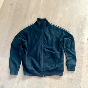 Svart Fred Perry ziptröja med vita ränder - Svart ziptröja från Fred Perry med vita ränder längs axlarna och det klassiska Fred Perry-logot på bröstet. Tröjan har hög krage, långa ärmar och två fickor framtill. Materialet är mjuk bomull och polyester, perfekt för chill dagar😎