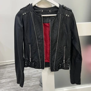 Svart skinnjacka från rockandblue - Svart äkta skinnjacka från rockandblue med coola silverfärgade dragkedjor och nitar. Jackan har röd insida, långärmade ärmar och detaljer vid axlar och midja. Snygg passform och klassisk bikerstil som lyfter din outfit. Använd en gång 